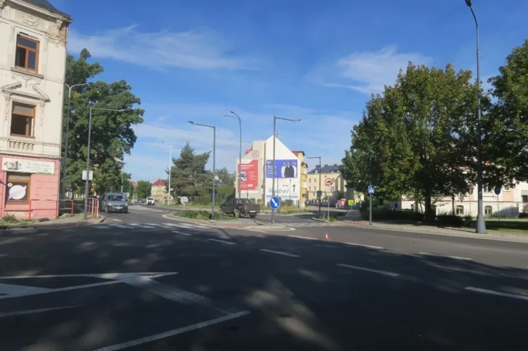Silnice na ulici Říční okruh v Krnově s dělicím ostrůvkem, přechodem pro chodce a dopravními značkami, v pozadí domy a stromy.