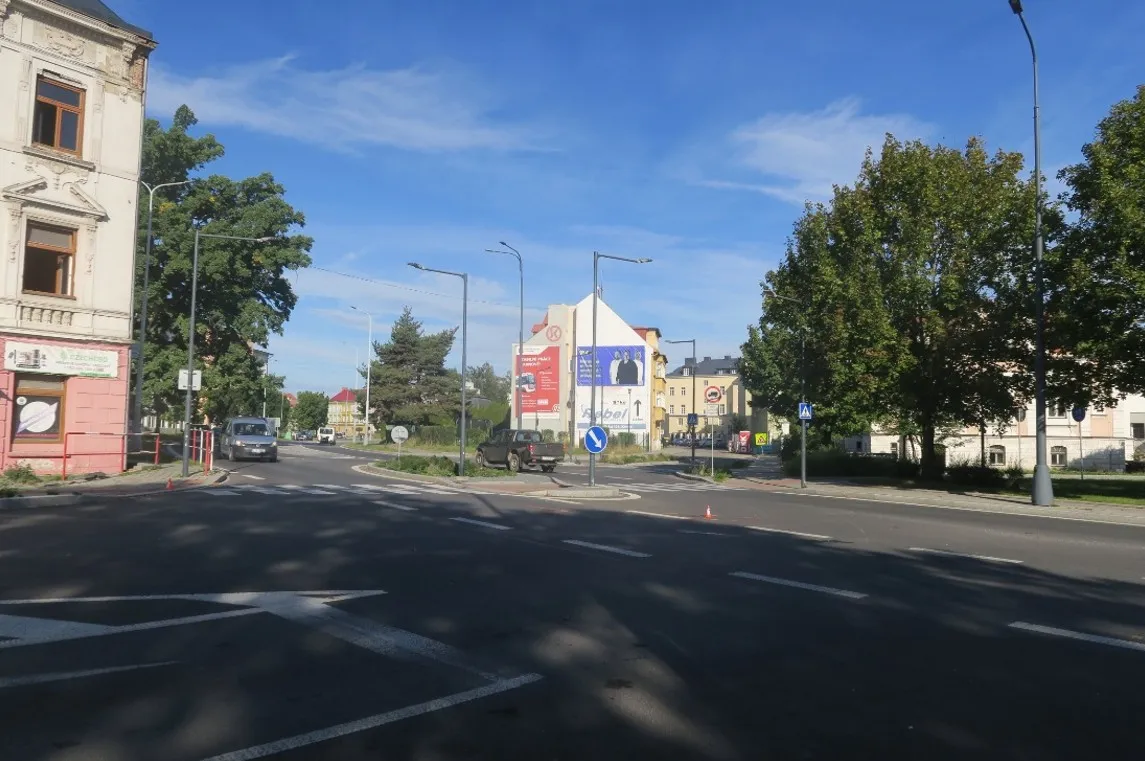 Silnice na ulici Říční okruh v Krnově s dělicím ostrůvkem, přechodem pro chodce a dopravními značkami, v pozadí domy a stromy.