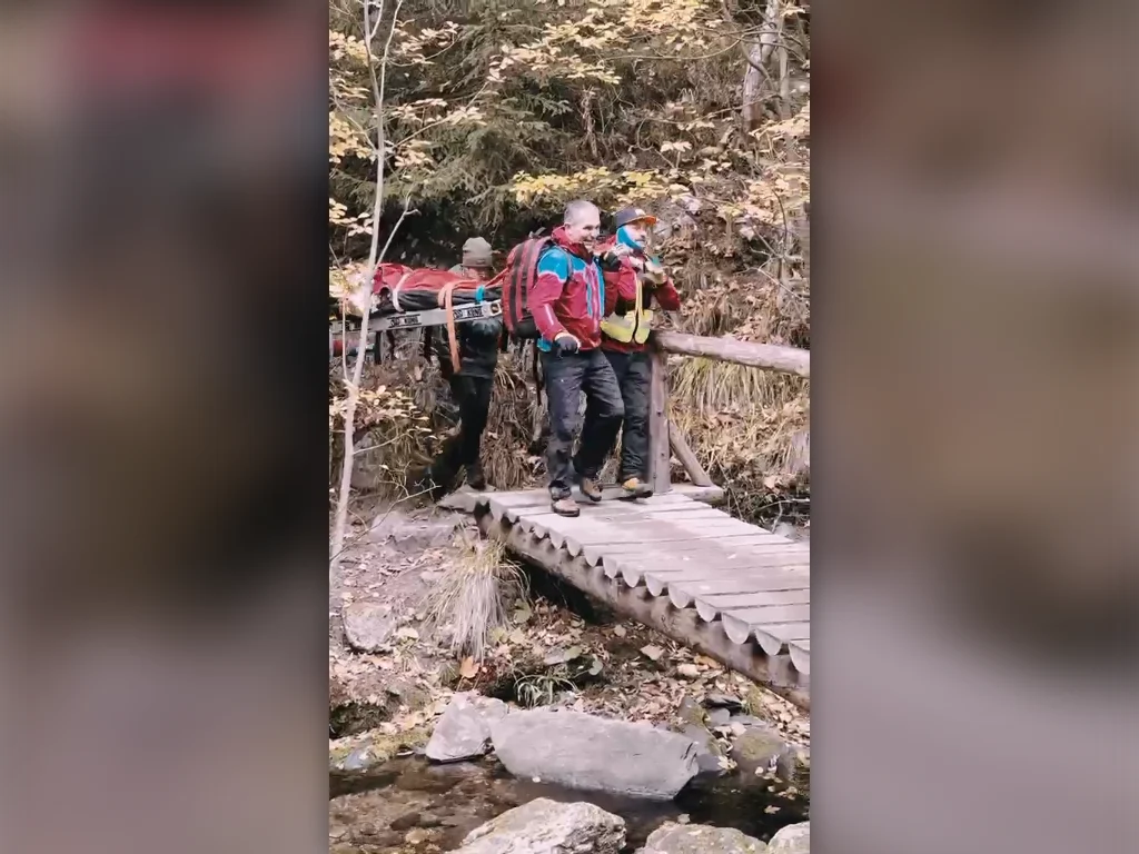 2026-10-19-ma-bila-opava-nahled (3) Záchranáři přenášejí nosítka se zraněnou turistkou přes dřevěnou lávku.